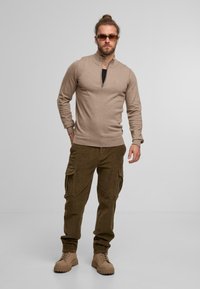 Beżowy sweter z zamkiem na szyi w połączeniu z oliwkowymi spodniami cargo z kordurowej tkaniny oraz brązowymi butami na sznurówki, z kieszeniami z klapami i ściągaczami.