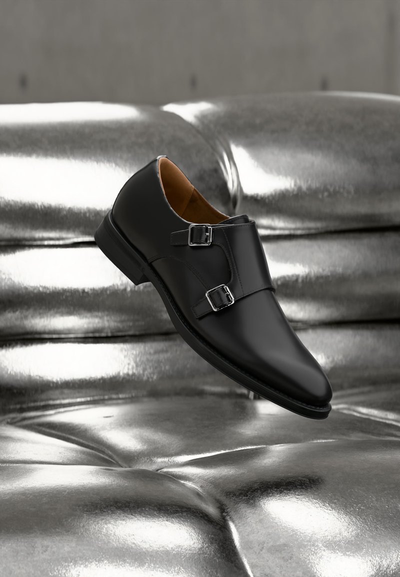 Chaussure noire en cuir à double boucle de moine avec des boucles argentées, présentée sur une surface capitonnée métallique argentée réfléchissante.