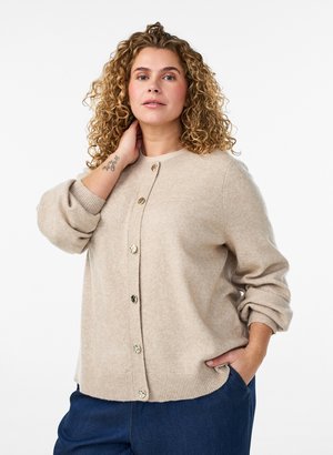 Woman with curly hair wearing beige buttoned cardigan and blue pants, posing with one hand on neck and the other in pocket.