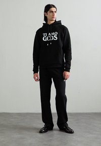 Sudadera negra con estampado "TI AMO GODS", combinada con pantalones negros. Tejido suave, corte relajado, cordones y diseño minimalista.