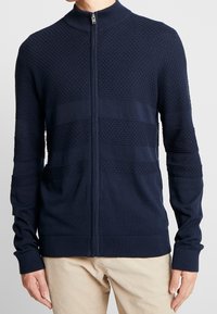 Marinblå zip-up tröja med ett texturerat mönster, med horisontella band, ribbade ärmslut och en hög krage av stickad material.