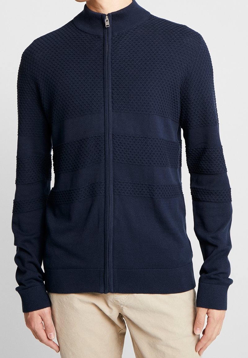 Marinblå zip-up tröja med ett texturerat mönster, med horisontella band, ribbade ärmslut och en hög krage av stickad material.