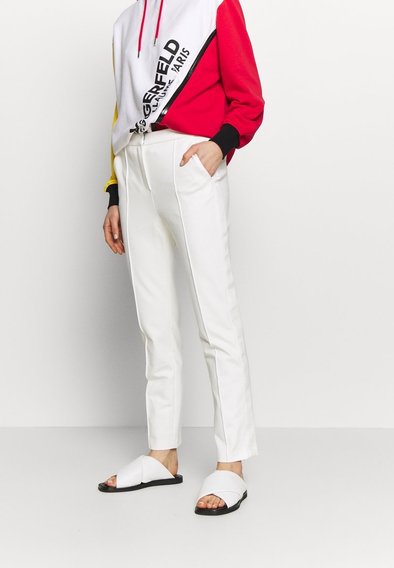 Weiße maßgeschneiderte Hosen mit Seitentaschen, kombiniert mit einem rot, schwarz und gelb farblich blockierten Hoodie. Getragen mit weißen crossover Rutsch-Sandalen.