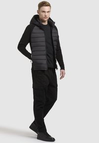 Gilet imbottito nero con cappuccio, abbinato a una maglietta a maniche lunghe nera e pantaloni cargo neri. Gli accessori includono sneaker nere.