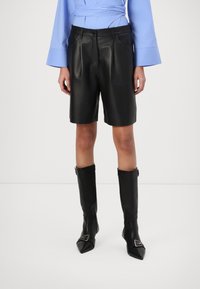 Shorts en cuir noir avec poches avant, conçus avec une texture lisse. Associés à des bottes en cuir noir montantes avec un accent de boucle.