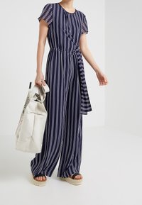 Jumpsuit a rayas azul marino con mangas cortas, cintura elástica y piernas anchas; combinado con un bolso de mano blanco y sandalias de plataforma.
