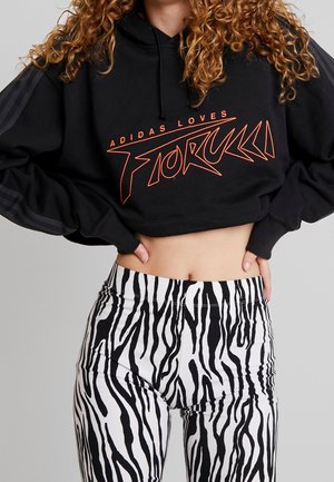 Osoba ubrana w czarną skróconą bluzę z kapturem z napisem "Adidas Loves Fiorucci" oraz wysokie spodnie w paski zebry, dłonie na biodrach.