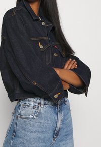 Tmavě modrá džínová bunda s krátkým střihem, stříbrnými knoflíky a oranžovým prošíváním. Kombinovaná s lehce opranými džínami s vysokým pasem.