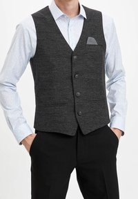 Uomo che indossa una camicia a righe azzurro chiaro, un gilet grigio scuro con taschino e pantaloni neri, con le mani nelle tasche.