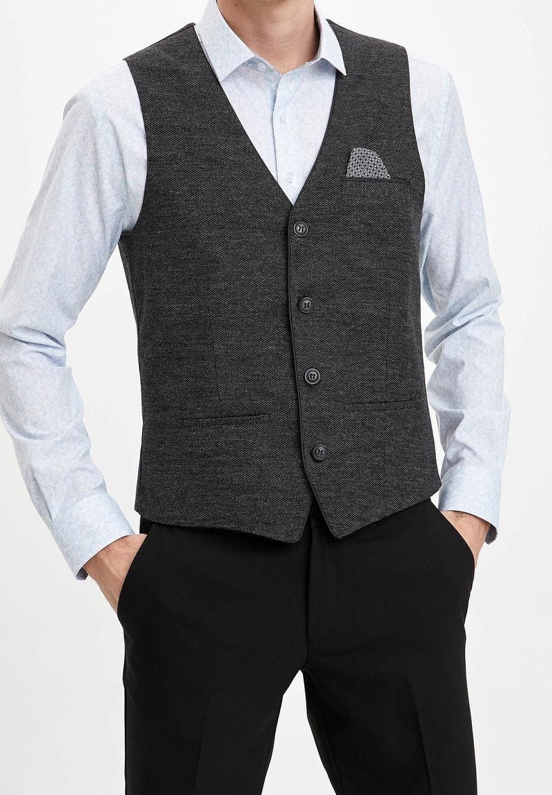 Uomo che indossa una camicia a righe azzurro chiaro, un gilet grigio scuro con taschino e pantaloni neri, con le mani nelle tasche.