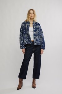 Tumesinise bandaanimustriga jakk, valge topp, tumedast denimist laiade säärtega püksid ja pruunid kontsaga saapad. Modell seisab neutraalse tausta ees.