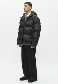 Chaqueta acolchada negra con capucha, cierre de cremallera y bolsillos laterales. Llevada sobre una camisa a rayas y pantalones anchos de pana negra. Zapatillas negras.