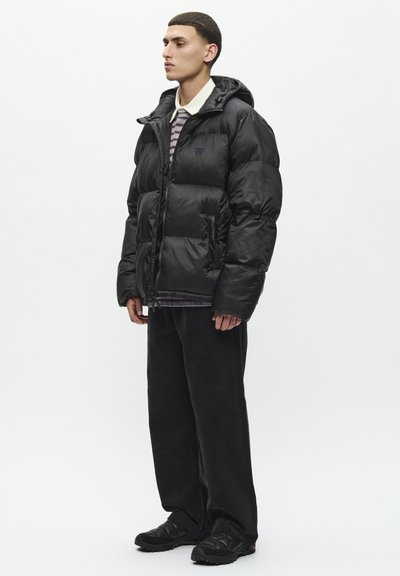 Veste en duvet noire avec capuche, fermeture éclair et poches latérales. Portée par-dessus une chemise rayée et un pantalon large en velours côtelé noir. Baskets noires.