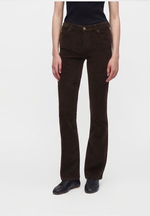 Personne portant un pantalon en velours côtelé brun foncé, un haut noir et des chaussures noires à lacets, debout devant un fond uni clair.