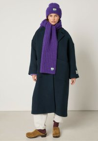 Mornarsko volneno plašč s prevelikim krojem, v kombinaciji z vijoličnim rebrastim šalom in klobukom, kremnimi trenirkami in rjavimi lesnimi copati.