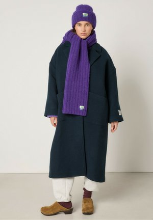 Navy wool overcoat with oversized fit, paired with a purple ribbed scarf and hat, cream sweatpants, and brown clogs.