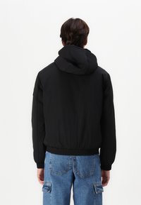 Osoba ubrana w czarną bluzę z kapturem i niebieskie jeansy bojówki, stojąca tyłem na tle gładkiej białej ściany.