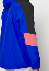 Modrá větrovka s černými a korálovými akcenty. Má kapuci, elastické manžety a hladkou, lehkou tkaninu.