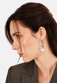 Srebrne kolczyki o płynnej, abstrakcyjnej formie z białą perłą. Kolczyki charakteryzują się wypolerowanym wykończeniem i unikalnym kształtem.