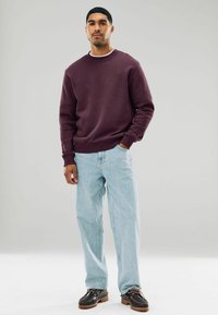 Sudadera burdeos con cuello redondo, puños y bajo acanalados, combinada con jeans azul claro y zapatos negros con acentos marrones.