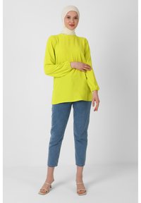 Lys gul langærmet top med samlede manchetter, parret med lyseblå jeans og beige hælede sandaler, sat mod en hvid baggrund.