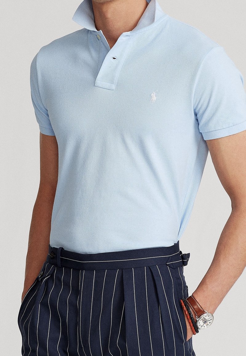 Homme portant un polo bleu clair à manches courtes avec col relevé, pantalon plissé à fines rayures bleu marine, montre et bracelets au poignet gauche.