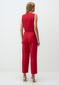 Red, sleeveless jumpsuit with a fitted bodice and wide-leg trousers. Features a smooth texture and clean lines, paired with cream heels.