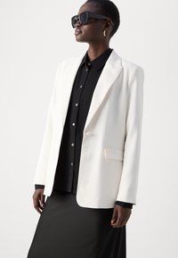 Weißer Blazer mit strukturierten Schultern, Ein-Knopf-Verschluss und Seitentaschen. Trägt man über einem schwarzen gerippten Shirt, kombiniert mit einem schwarzen Rock.