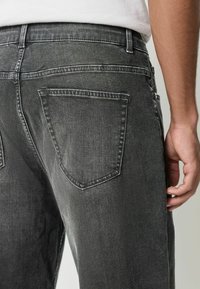 Ciemnoszare dżinsy o klasycznym pięciokieszeniowym kroju. Tkanina ma lekki połysk, widoczne szwy i teksturowane wykończenie.