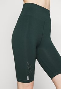 Zbliżenie na osobę w ciemnozielonych, wysokich szortach sportowych do kolan z oddychającymi detalami z siateczki oraz małą metką B.DRY.