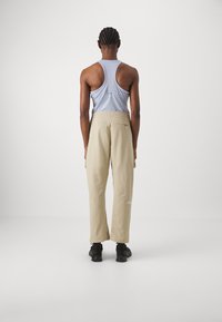 Person med flätat hår som bär en ljusblå racerback-top, beige cargobyxor och svarta skor, stående mot en vit bakgrund.