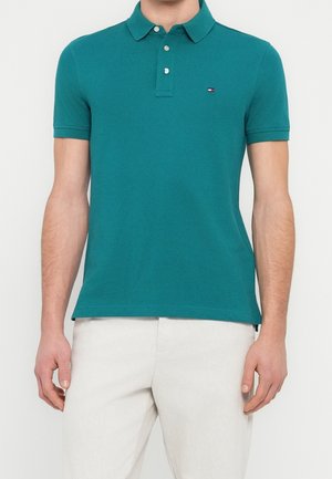 Homme portant un polo manches courtes couleur sarcelle avec un petit logo sur la poitrine et un pantalon écru sur fond uni.