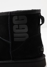 Černá semišová kotníková bota s jemnou texturou, se výrazným černým logem "UGG" na boku a pevnou černou gumovou podrážkou.