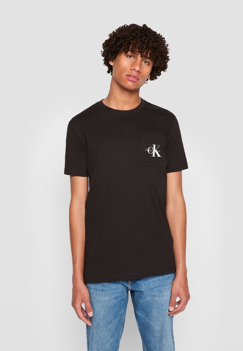 Junger Mann mit lockigem Haar, der ein schwarzes Calvin Klein T-Shirt und blaue Jeans trägt, vor einem schlichten hellgrauen Hintergrund stehend.