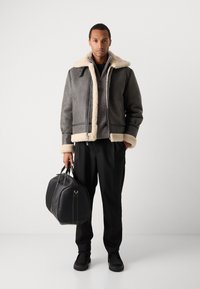 Γκρι τζάκετ με επένδυση από γούνα, με μπροστινό φερμουάρ, σε συνδυασμό με σκούρα παντελόνια και μαύρη δερμάτινη τσάντα duffle, σε ουδέτερο φόντο.