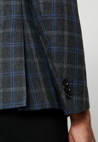 Grau karierter Blazer mit blauen Akzenten, aus strukturiertem Stoff, mit einem strukturierten Schulterdesign und drei schwarzen Knöpfen am Ärmel.