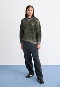 Pulover texturat, verde închis, cu decolteu la baza gâtului, asortat cu pantaloni negri largi și adidași negri cu dungi albe. Ochelari clare purtați.