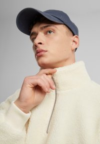 Kremowy polarowy pulower z wysokim kołnierzem i zamkiem. Granatowa czapka modela i ręka na kołnierzu podkreślają miękką teksturę i nowoczesny design.
