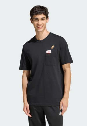 Uomo sorridente con t-shirt nera con grafica di pallacanestro e canestro sopra la tasca sul petto, indossa pantaloni neri Adidas con logo sulla coscia sinistra.