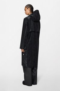 Μαύρο, μακρύ αδιάβροχο με υφασμέν.pattern μεταλλικής υφής, κουκούλα και πλαϊνές τσέπες, σε συνδυασμό με φαρδιά μαύρα παντελόνια και επίπεδα παπούτσια.