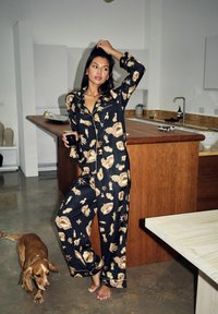 Set de pijamale negre cu modele aurii de mâini și ochi, cămașă cu nasturi și pantaloni largi. Blat de lemn și un câine maro în apropiere.