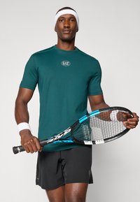 Camisa atlética de manga curta em teal com tecnologia de absorção de humidade e logótipo, combinada com calções pretos. O modelo segura uma raquete de ténis com detalhes em azul.