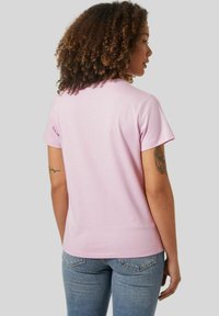Camiseta de algodón rosa claro con mangas cortas, cuello redondo y una textura suave, combinada con jeans azules. La vista posterior no muestra detalles gráficos.