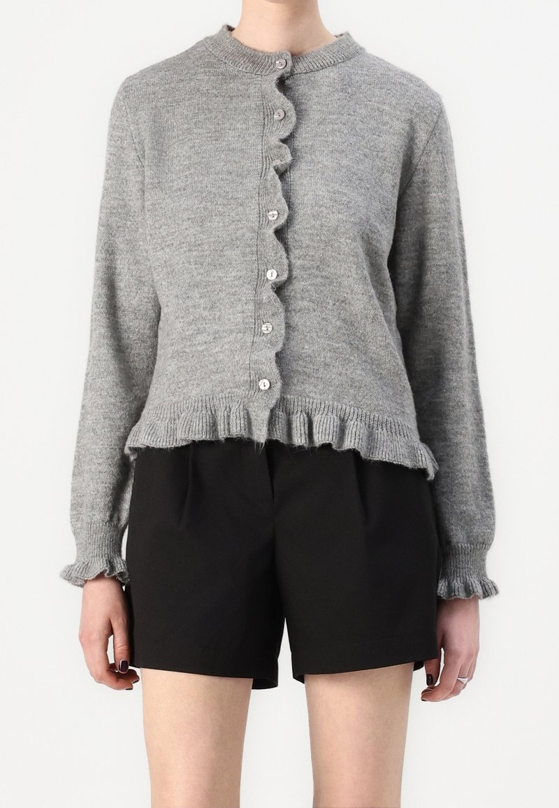 Cardigan gris en maille avec bords à volants et boutons, associé à un short taille haute noir porté par une personne debout sur un fond uni.
