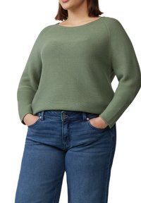 Groene geribbelde trui met een ronde halslijn en lange mouwen, gecombineerd met blauwe jeans. De trui heeft een ontspannen pasvorm en een gestructureerde stof.