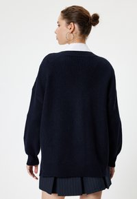 Tummansininen neulepaita pitkillä hihoilla, ribbikuviointi ja löysä istuvuus. Takana kauluksen korkeus ja se on yhdistetty laskostettuihin hameisiin.