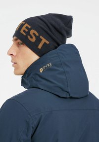 Chaqueta impermeable azul con un cuello alto y un logo en la parte posterior, llevada con un gorro oscuro que presenta la palabra "TEST" en letras doradas.