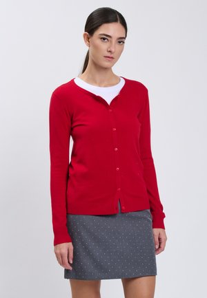 Kvinde iført en rød knappet cardigan over en hvid top og et gråt skørt med små hvide prikker, stående mod en ensfarvet baggrund.