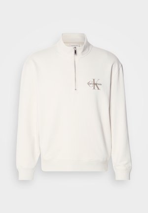 Bílý svetr s polo zipem a šedým logem na levé straně hrudi, s žebrovanými manžetami a spodním lemem. Měkká textury materiálu.