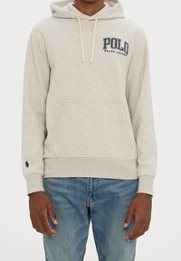Světlé šedé mikina s kapucí s přední kapsou, která má béžové šňůrky a námořnicky modré vyšívané logo "Polo Ralph Lauren" na hrudi.